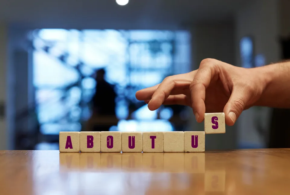 A hand placing the final wooden block to complete the words About Us on a table.